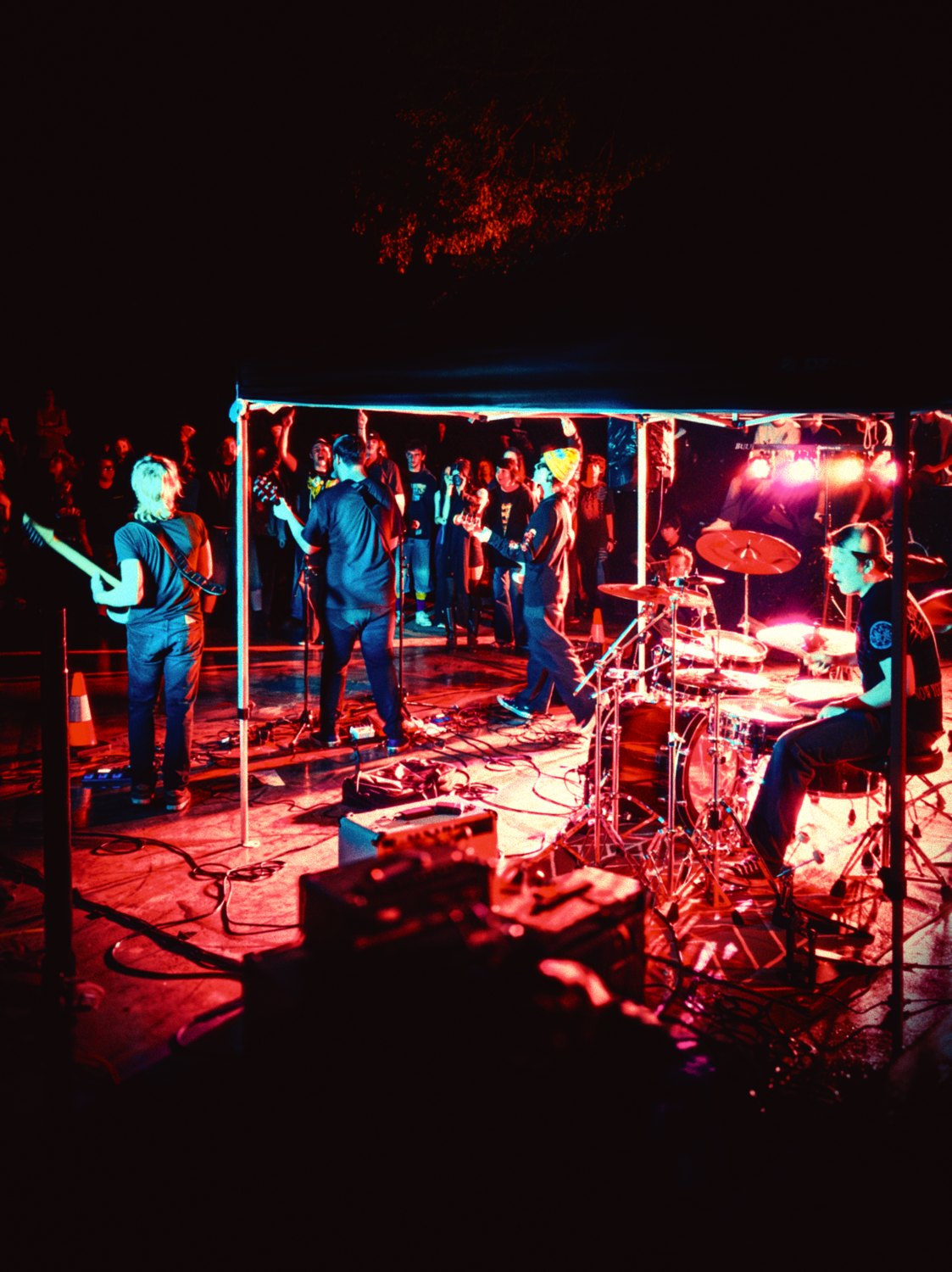 Delphic Afterparty playing under a gazebo in red stage light, the wet concrete throwing the light back, the crowd close in